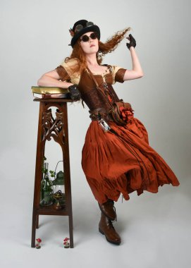 Uzun, yürüyen Steampunk Botanikçisi, çılgın bilim adamı, silindir şapkalı ve gözlüklü kadın model. Kızıl Saçlı Fantezi Karakteri Poz Beyaz Stüdyo Arkaplanı hakkında akademik kitaplar tutuyor