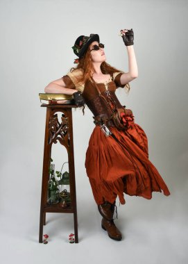 Uzun, yürüyen Steampunk Botanikçisi, çılgın bilim adamı, silindir şapkalı ve gözlüklü kadın model. Kızıl Saçlı Fantezi Karakteri Poz Beyaz Stüdyo Arkaplanı hakkında akademik kitaplar tutuyor