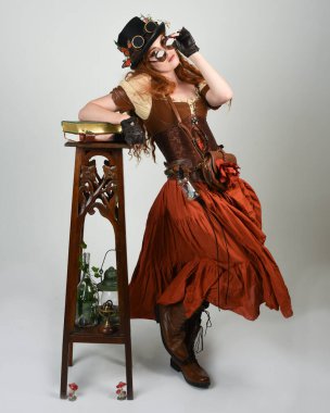 Uzun, yürüyen Steampunk Botanikçisi, çılgın bilim adamı, silindir şapkalı ve gözlüklü kadın model. Kızıl Saçlı Fantezi Karakteri Poz Beyaz Stüdyo Arkaplanı hakkında akademik kitaplar tutuyor