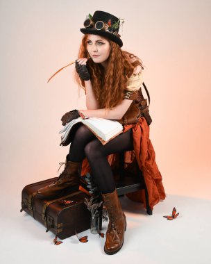 Steampunk Viktorya Dönemi kostümlü, silindir şapkalı ve gözlüklü kadın model. Kızıl Saçlı Fantezi Karakteri oturmuş, Beyaz Stüdyo Arkaplan kitabını yazıyor.