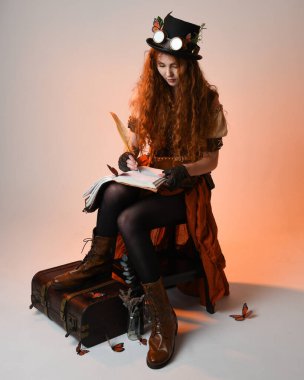 Steampunk Viktorya Dönemi kostümlü, silindir şapkalı ve gözlüklü kadın model. Kızıl Saçlı Fantezi Karakteri oturmuş, Beyaz Stüdyo Arkaplan kitabını yazıyor.