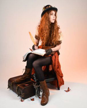 Steampunk Viktorya Dönemi kostümlü, silindir şapkalı ve gözlüklü kadın model. Kızıl Saçlı Fantezi Karakteri oturmuş, Beyaz Stüdyo Arkaplan kitabını yazıyor.