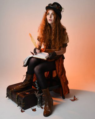 Steampunk Viktorya Dönemi kostümlü, silindir şapkalı ve gözlüklü kadın model. Kızıl Saçlı Fantezi Karakteri oturmuş, Beyaz Stüdyo Arkaplan kitabını yazıyor.