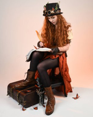 Steampunk Viktorya Dönemi kostümlü, silindir şapkalı ve gözlüklü kadın model. Kızıl Saçlı Fantezi Karakteri oturmuş, Beyaz Stüdyo Arkaplan kitabını yazıyor.