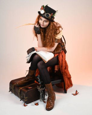 Steampunk Viktorya Dönemi kostümlü, silindir şapkalı ve gözlüklü kadın model. Kızıl Saçlı Fantezi Karakteri oturmuş, Beyaz Stüdyo Arkaplan kitabını yazıyor.