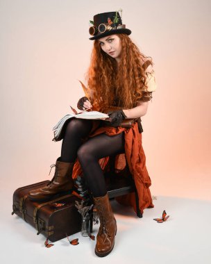 Steampunk Viktorya Dönemi kostümlü, silindir şapkalı ve gözlüklü kadın model. Kızıl Saçlı Fantezi Karakteri oturmuş, Beyaz Stüdyo Arkaplan kitabını yazıyor.