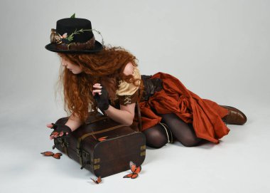 Steampunk Botanikçisi, deli bilim adamı, silindir şapkalı ve gözlüklü kadın model. Kızıl Saçlı Fantezi Karakteri Beyaz Stüdyo Arkaplanda Kelebeklerle Poz Veriyor