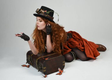 Steampunk Botanikçisi, deli bilim adamı, silindir şapkalı ve gözlüklü kadın model. Kızıl Saçlı Fantezi Karakteri Beyaz Stüdyo Arkaplanda Kelebeklerle Poz Veriyor