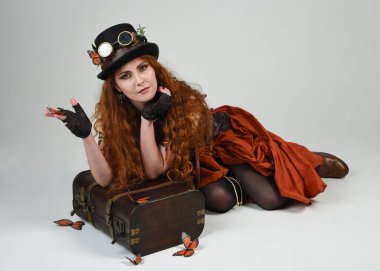 Steampunk Botanikçisi, deli bilim adamı, silindir şapkalı ve gözlüklü kadın model. Kızıl Saçlı Fantezi Karakteri Beyaz Stüdyo Arkaplanda Kelebeklerle Poz Veriyor