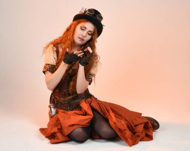 Steampunk Viktorya Dönemi kostümlü, silindir şapkalı ve gözlüklü kadın model. Kızıl Saçlı Fantezi Karakteri. Beyaz Stüdyo Arkaplanı 'nda diz çökmüş poz