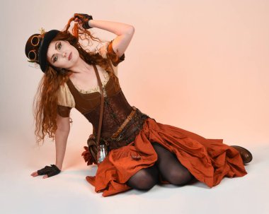 Steampunk Viktorya Dönemi kostümlü, silindir şapkalı ve gözlüklü kadın model. Kızıl Saçlı Fantezi Karakteri. Beyaz Stüdyo Arkaplanı 'nda diz çökmüş poz