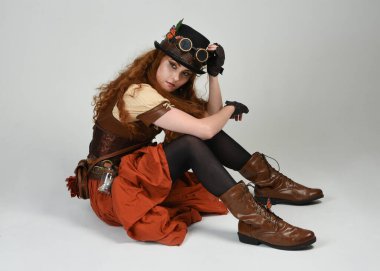 Steampunk Viktorya Dönemi kostümlü, silindir şapkalı ve gözlüklü kadın model. Kızıl Saçlı Fantezi Karakteri. Beyaz Stüdyo Arkaplanı 'nda diz çökmüş poz
