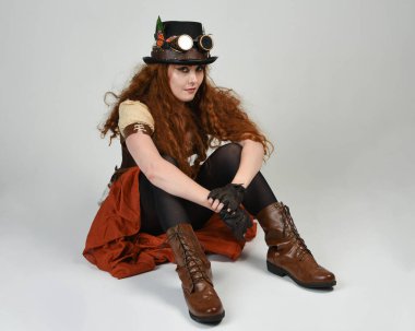 Steampunk Viktorya Dönemi kostümlü, silindir şapkalı ve gözlüklü kadın model. Kızıl Saçlı Fantezi Karakteri. Beyaz Stüdyo Arkaplanı 'nda diz çökmüş poz