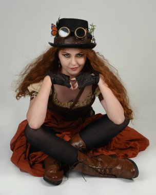 Steampunk Viktorya Dönemi kostümlü, silindir şapkalı ve gözlüklü kadın model. Kızıl Saçlı Fantezi Karakteri. Beyaz Stüdyo Arkaplanı 'nda diz çökmüş poz