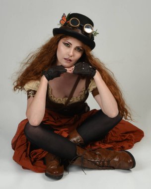 Steampunk Viktorya Dönemi kostümlü, silindir şapkalı ve gözlüklü kadın model. Kızıl Saçlı Fantezi Karakteri. Beyaz Stüdyo Arkaplanı 'nda diz çökmüş poz