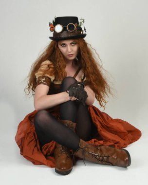 Steampunk Viktorya Dönemi kostümlü, silindir şapkalı ve gözlüklü kadın model. Kızıl Saçlı Fantezi Karakteri. Beyaz Stüdyo Arkaplanı 'nda diz çökmüş poz