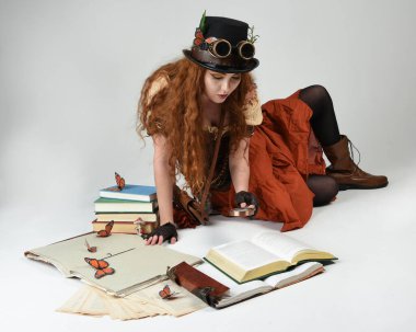 Steampunk Viktorya Dönemi kostümlü, silindir şapkalı ve gözlüklü kadın model. Kızıl Saçlı Fantezi Karakteri oturmuş poz kitabı okuyor, Beyaz Stüdyo Arkaplan