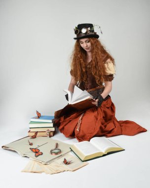 Steampunk Viktorya Dönemi kostümlü, silindir şapkalı ve gözlüklü kadın model. Kızıl Saçlı Fantezi Karakteri oturmuş poz kitabı okuyor, Beyaz Stüdyo Arkaplan