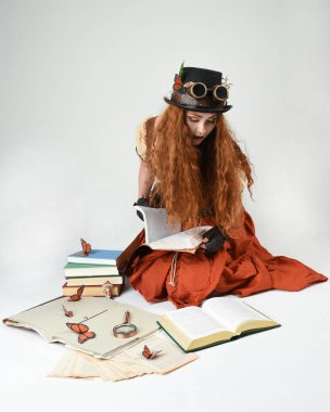Steampunk Viktorya Dönemi kostümlü, silindir şapkalı ve gözlüklü kadın model. Kızıl Saçlı Fantezi Karakteri oturmuş poz kitabı okuyor, Beyaz Stüdyo Arkaplan