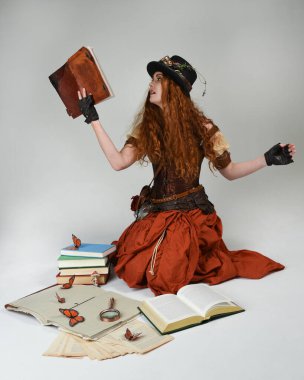 Steampunk Viktorya Dönemi kostümlü, silindir şapkalı ve gözlüklü kadın model. Kızıl Saçlı Fantezi Karakteri oturmuş poz kitabı okuyor, Beyaz Stüdyo Arkaplan