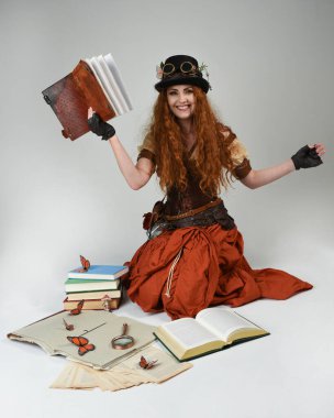 Steampunk Viktorya Dönemi kostümlü, silindir şapkalı ve gözlüklü kadın model. Kızıl Saçlı Fantezi Karakteri oturmuş poz kitabı okuyor, Beyaz Stüdyo Arkaplan