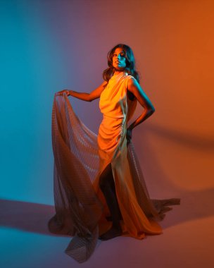 Fantezi bir taç takan kadın bir modelin tam boy portresi, sarı elbise ve akıcı kumaş pelerin. Sıcak altın günbatımı ışıklarıyla stüdyo arka planında yürüyüş pozu