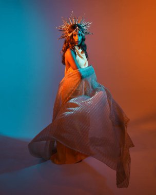 Fantezi bir taç takan kadın bir modelin tam boy portresi, sarı elbise ve akıcı kumaş pelerin. Sıcak altın günbatımı ışıklarıyla stüdyo arka planında yürüyüş pozu