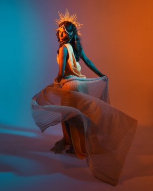 Fantezi bir taç takan kadın bir modelin tam boy portresi, sarı elbise ve akıcı kumaş pelerin. Sıcak altın günbatımı ışıklarıyla stüdyo arka planında yürüyüş pozu