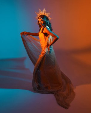 Fantezi bir taç takan kadın bir modelin tam boy portresi, sarı elbise ve akıcı kumaş pelerin. Sıcak altın günbatımı ışıklarıyla stüdyo arka planında yürüyüş pozu