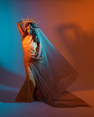 Fantezi bir taç takan kadın bir modelin tam boy portresi, sarı elbise ve akıcı kumaş pelerin. Sıcak altın günbatımı ışıklarıyla stüdyo arka planında yürüyüş pozu