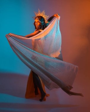 Fantezi bir taç takan kadın bir modelin tam boy portresi, sarı elbise ve akıcı kumaş pelerin. Sıcak altın günbatımı ışıklarıyla stüdyo arka planında yürüyüş pozu