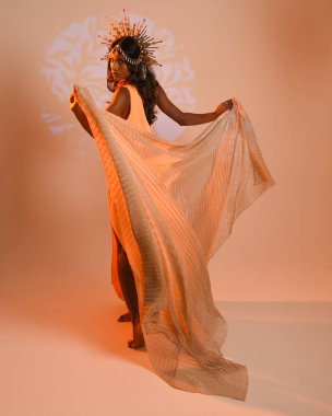 Fantezi bir taç takan kadın bir modelin tam boy portresi, sarı elbise ve akıcı kumaş pelerin. Stüdyonun arka planında yürüme pozu