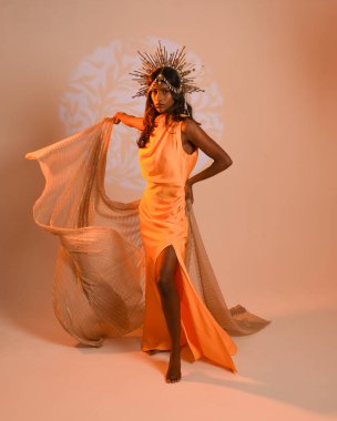 Fantezi bir taç takan kadın bir modelin tam boy portresi, sarı elbise ve akıcı kumaş pelerin. Stüdyonun arka planında yürüme pozu