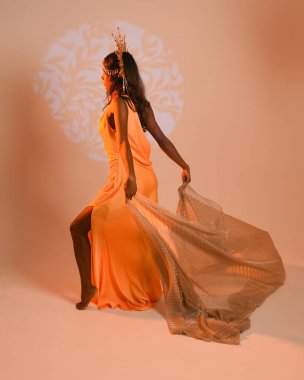 Fantezi bir taç takan kadın bir modelin tam boy portresi, sarı elbise ve akıcı kumaş pelerin. Stüdyonun arka planında yürüme pozu