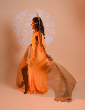 Fantezi bir taç takan kadın bir modelin tam boy portresi, sarı elbise ve akıcı kumaş pelerin. Stüdyonun arka planında yürüme pozu