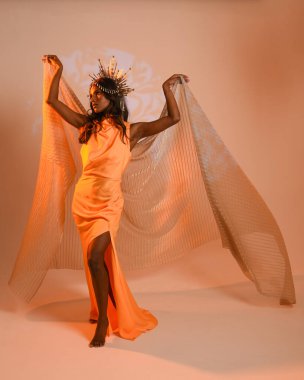 Fantezi bir taç takan kadın bir modelin tam boy portresi, sarı elbise ve akıcı kumaş pelerin. Stüdyonun arka planında yürüme pozu