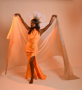 Fantezi bir taç takan kadın bir modelin tam boy portresi, sarı elbise ve akıcı kumaş pelerin. Stüdyonun arka planında yürüme pozu