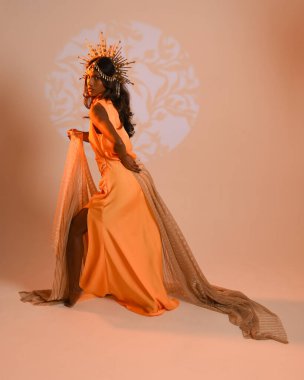 Fantezi bir taç takan kadın bir modelin tam boy portresi, sarı elbise ve akıcı kumaş pelerin. Stüdyonun arka planında yürüme pozu