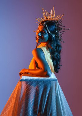 Güzel esmer bir kadının portresi. Fantezi tacını takıyor ve göz alıcı sarı bir balo elbisesi giyiyor. Sinematik renkli ışıklandırmayla stüdyonun arka planında zarif bir poz..