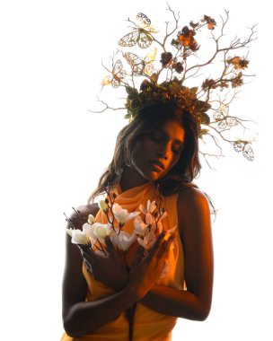 Arka planda, altın sarısı bir ışıkla parıldayan, çiçekli bir taç takan, elinde çiçekler tutan, yumuşak, ruhani bir fantezi tanrıçası fantezi karakterini anımsatan bir kadının siluet portresi.