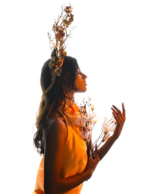 Arka planda, altın sarısı bir ışıkla parıldayan, çiçekli bir taç takan, elinde çiçekler tutan, yumuşak, ruhani bir fantezi tanrıçası fantezi karakterini anımsatan bir kadının siluet portresi.