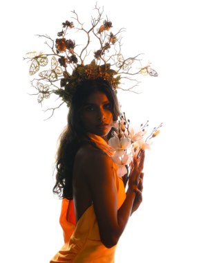 Arka planda, altın sarısı bir ışıkla parıldayan, çiçekli bir taç takan, elinde çiçekler tutan, yumuşak, ruhani bir fantezi tanrıçası fantezi karakterini anımsatan bir kadının siluet portresi.