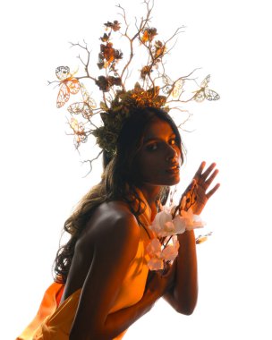 Arka planda, altın sarısı bir ışıkla parıldayan, çiçekli bir taç takan, elinde çiçekler tutan, yumuşak, ruhani bir fantezi tanrıçası fantezi karakterini anımsatan bir kadının siluet portresi.