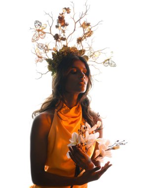 Arka planda, altın sarısı bir ışıkla parıldayan, çiçekli bir taç takan, elinde çiçekler tutan, yumuşak, ruhani bir fantezi tanrıçası fantezi karakterini anımsatan bir kadının siluet portresi.