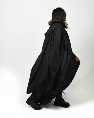 Siyah deri kostüm ve pelerin giyen, yürüyüp giden esmer kadının vücut portresi. Beyaz stüdyo arka planında izole edilmiş güçlü gotik fantezi karakteri.