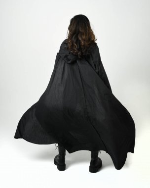 Siyah deri kostüm ve pelerin giyen, yürüyüp giden esmer kadının vücut portresi. Beyaz stüdyo arka planında izole edilmiş güçlü gotik fantezi karakteri.