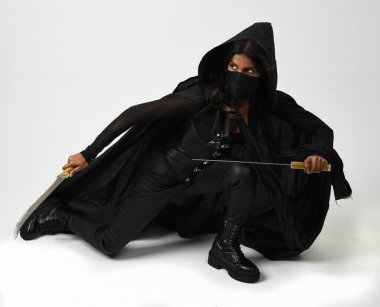 Siyah deri kostüm ve pelerin giyen, elinde kılıç tutan esmer kadının vücut portresi. Diz çökmüş güçlü gotik fantezi karakteri. Beyaz stüdyo arka planında izole