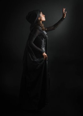  Siyah pelerinli gizemli kapüşonlu kadın portresi, gotik fantezi ya da distopist karakter. Sihirli el hareketleriyle poz vermek. Huysuz gölgelerle aydınlatılmış, karanlık izole edilmiş arka planda stüdyo sahnesi