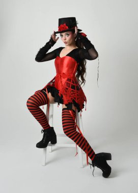 Burlesque sirk sanatçısı, Cadılar Bayramı soytarısı kostümlü fantezi kadın model silindir şapkalı, kırmızı gotik korse. Sandalyeye oturmuş tiyatro pozu, izole edilmiş beyaz stüdyo arka planı..