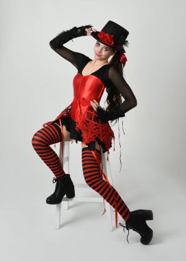 Burlesque sirk sanatçısı, Cadılar Bayramı soytarısı kostümlü fantezi kadın model silindir şapkalı, kırmızı gotik korse. Sandalyeye oturmuş tiyatro pozu, izole edilmiş beyaz stüdyo arka planı..